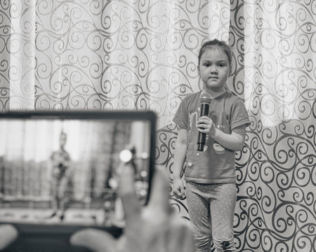 Cute little Asian girl looking at camera holding microphone at home concert. Hands holding tablet for taking video, in unfocus. lifestyle concept. Black and white snapshot. selective focusの写真素材