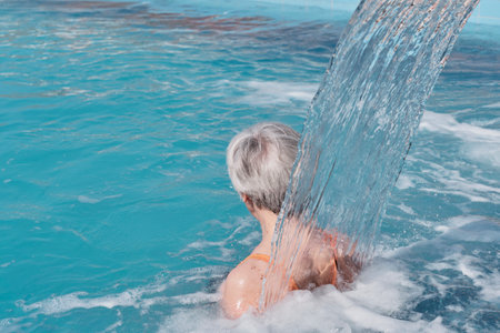 Senior woman with gray hair under flow of water falling from above. rear view. Hydromassage in outdoor thermal swimming pool. Active elderly concept.の写真素材