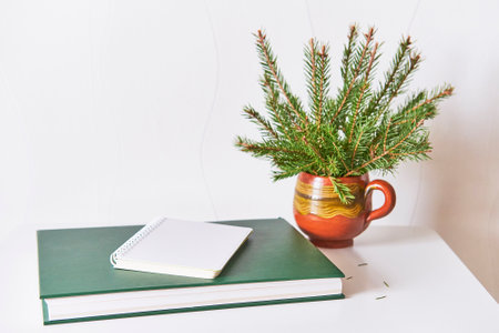 Notepad with blank sheet, lying on green book, bouquet of christmas tree branches in clay mugの写真素材