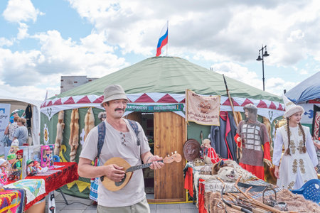 Irbit, Russia - August 11, 2023: Senior Asian man jokingly playing national string instrument against backdrop of national Bashkir courtyard in form of decorated tent. Traditional Irbit fair.のeditorial素材