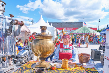 Irbit, Russia - August 11, 2023: Portrait of senior white woman in festive folk clothes, looking at camera, standing at table, on which big old metal samovar and kitchenware.Traditional Irbit fairのeditorial素材