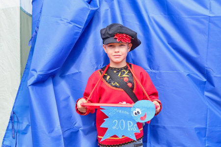 Irbit, Russia - August 11, 2023: Portrait of young boy as peddler of small items on traditional Irbit fair, in stylized Russian national red shirt with embroidery and cap with handmade flower.のeditorial素材