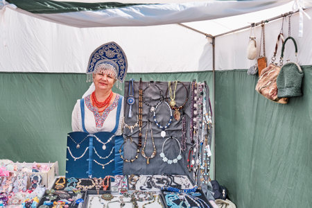 Irbit, Russia - August 11, 2023: Senior Caucasian smiling saleswoman in Russian national clothes and embroidered kokoshnik. Selling costume jewelry and accessories in trade tent at traditional fair.のeditorial素材