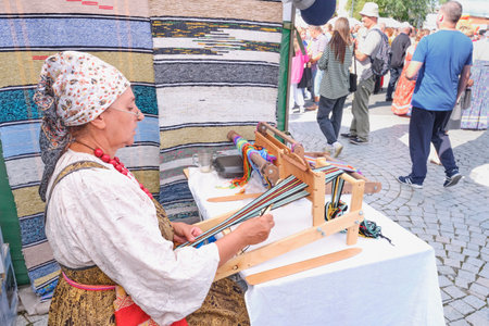 Irbit, Russia - August 11, 2023: Senior white woman at decorated tent weaving colorful striped belt on hand loom. She wearing traditional Russian clothes. Events of traditional Irbit fair.のeditorial素材