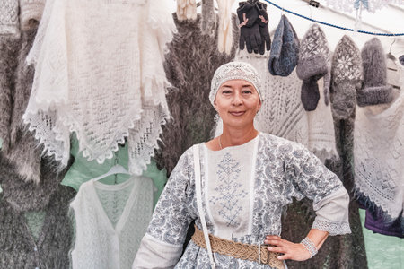 Irbit, Russia - August 11, 2023: Pleasant senior white woman hanging white fluffy shawl on hanger. She selling warm knitted items made of wool and fluff at traditional fair.のeditorial素材