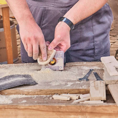 Male hands sanding edges of wooden little heart using small electric machine tool. Making souvenir,...