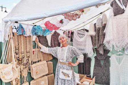 Irbit, Russia - August 11, 2023: Portrait of nice senior white woman, looking at camera, selling warm knitted items made of wool and fluff and crochet accessories at traditional fair.のeditorial素材