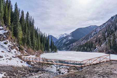 Scenic Kolsai lake ice-covered, surrounded by hillsides with green spruces and snow. Northern Tien Shan, Kolsai gorge, Kazakhstan. Spring landscape with pedestrian wooden bridgeの写真素材