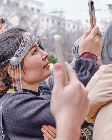 Almaty, Kazakhstan - March 21, 2024: Cute Asian woman wearing traditional Kazakh metal headband, filming concert on her phone in crowd during celebration of spring holiday of Nowruz.のeditorial素材
