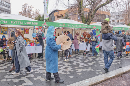 Almaty, Kazakhstan - March 20, 2024: People walking at crafts fair along pedestrian street during spring holiday of Nowruz. Man in Kazakh clothes beats tambourine. Cityscape on rainy dayのeditorial素材