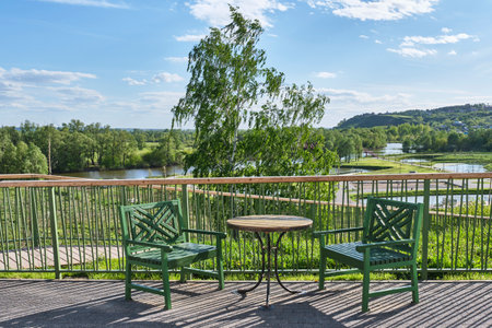 Round table and two wooden chairs on path near fence, place to relax. View of Shishkinskiye Ponds Culture and Recreation Park and Toyma River, Yelabuga, Russia. Spring cityscape.の写真素材