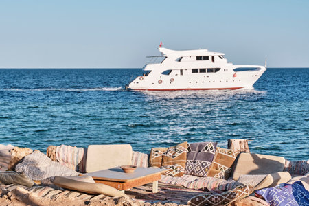 Cozy outdoor seating with colorful patterned pillows overlooks a serene sea, with a luxury yacht sailing by in the background. Perfect setting for relaxation and seaside leisure.の写真素材