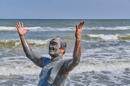 Senior man covered in therapeutic mud on beach of sea enjoys life with arms raised in sunny day. Sea of Azov, Temryuksky District, Russia.の写真素材