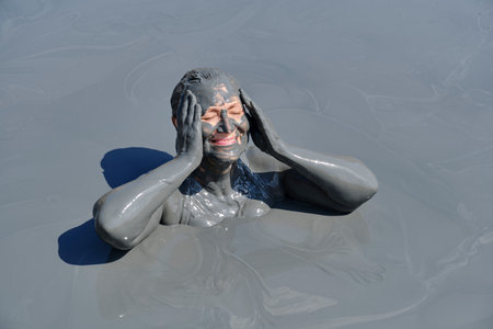 Senior woman enjoying mud bath with her face and body covered in therapeutic gray mud, smiling and...