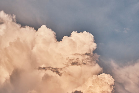 Close-up of fluffy white and beige clouds illuminated by warm sunset light against clear blue sky. Peaceful and dramatic beauty of nature's sky textures.の写真素材