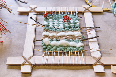 Handmade weaving project on wooden frame, featuring green, white, and brown yarn with natural elements like twigs and red berries for rustic, artistic touch.の写真素材