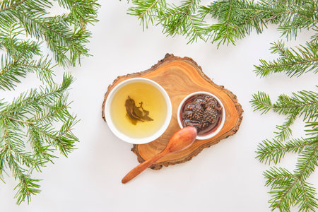 Cup of herbal tea and bowl of pine cone jam on wooden serving board, wooden spoon. Top view. Scene is surrounded by fresh fir branches on white background. Natural and cozy aesthetic.の写真素材