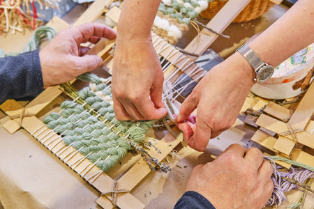 Close-up of hands weaving small tapestry on handmade wooden frame using green and white threads with...