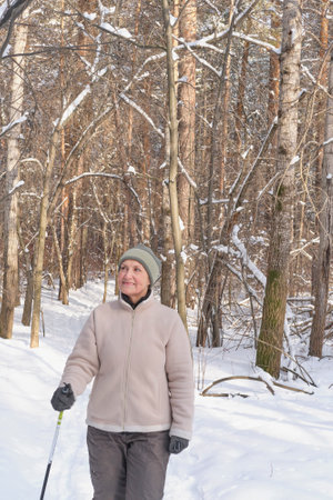 Senior woman cross-country skiing through snowy forest. She smiles surrounded by snow-covered trees. Sunlight streams through the branches. Tranquil outdoor atmosphere.の写真素材