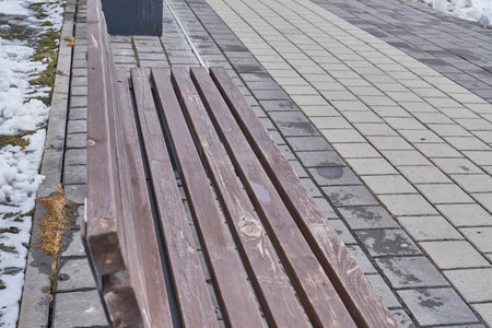 Weathered wooden bench in outdoor urban environment, surrounded by wet pavement and patches of melting snow. Scene of sense of solitude, cold weather, and seasonal transition.の写真素材