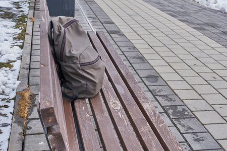 Worn backpack rests on wooden bench in urban outdoor setting. Surrounding pavement is wet, with patches of melting snow nearby. Scene of sense of solitude, mystery, or forgotten moment.の写真素材