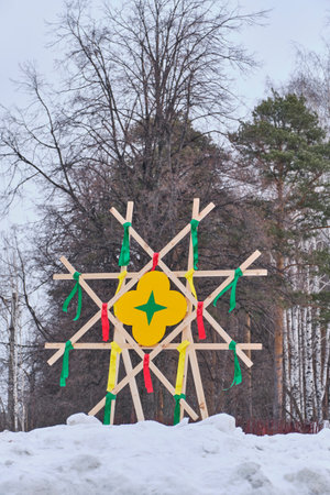 Decorative wooden structure with colorful ribbons and yellow center symbol set in snowy park during Maslenitsa celebration, surrounded by coniferous and bare trees.の写真素材