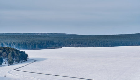 Curved ice skating track on frozen surface of Sysert city pond in winter, surrounded by snowy forest and hills under cloudy sky, Russiaの写真素材