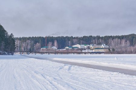 Ice skating trail on frozen pond in Sysert, Russia. Pedestrian bridge leads to snow-covered village Luch with forest backdrop. Peaceful winter rural landscape under clear sky.の写真素材