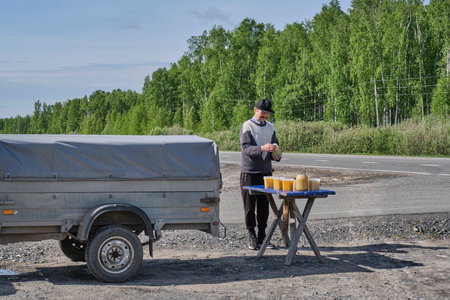 A beekeeper sells agricultural products, bee products, to passing travelers. Next to a cargo trailer there is a wooden table with several jars of honey against the backdrop of a road landscape.の写真素材