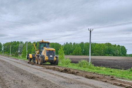 Motor grader performing work on leveling dirt road in countryside. Rear view. Rural landscape on...