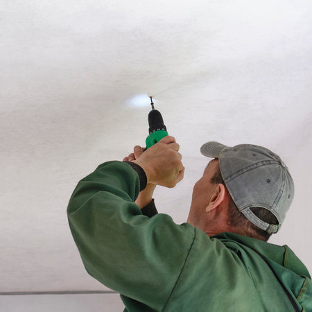 Worker wearing green jacket and gray cap uses cordless electric screwdriver to install or repair ceiling indoors, bottom view.の写真素材
