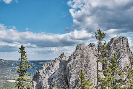 Dramatic granite rock formations with scattered pines under a blue sky in Taganay National Park, Southern Urals, Russia. A stone river lies below, framed by forested hills.の写真素材