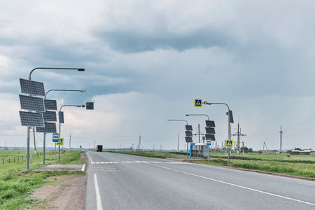 Empty countryside road with pedestrian crossings and bus stops equipped with solar-powered street lamps and traffic signs, under overcast sky in rural area.の写真素材