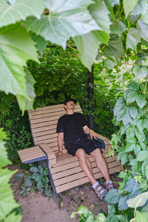 Man relaxing on a wooden bench under dense green foliage in a shaded park area. Peaceful moment of rest in nature during summer.の写真素材