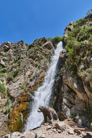 Scenic waterfall cascading down rocky cliffs in Turgen Gorge, Kazakhstan, with a realistic bear sculpture at the base. Surrounded by rugged terrain and lush greenery under clear blue sky.の写真素材