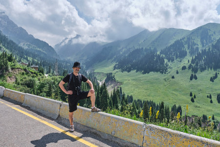 Male hiker in casual outfit standing on roadside barrier, posing lush green mountain valley under cloudy sky. Small Almaty Gorge, Shymbulak, Kazakhstan. Summer travel and adventureの写真素材