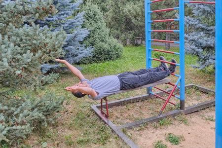 A senior man in casual sportswear exercises on a press bench in an outdoor sports complex among...