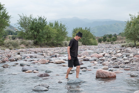 A senior man in black clothes and a Panama hat crossing a shallow rocky mountain river, surrounded...