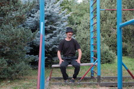 Senior man in casual clothing sitting on workout bench at outdoor gym, surrounded by green trees. Relaxing moment in park fitness area.の写真素材