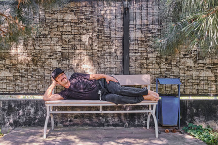 Senior man in casual clothes reclining on bench in shaded park area, resting barefoot with relaxed pose. Outdoor leisure and relaxation concept.の写真素材