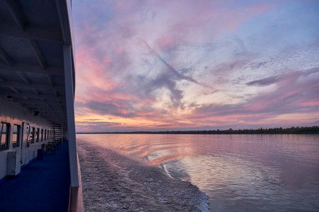 Tranquil view from river cruise ship deck during colorful sunset, with calm water, rippling wake, and soft pink and purple clouds reflecting on surfaceの写真素材