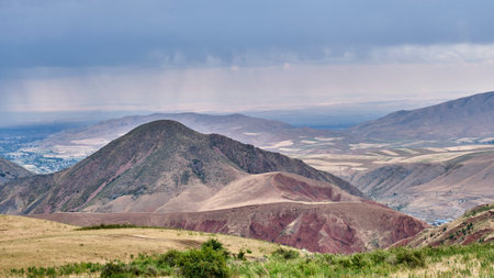 Majestic panoramic view of the rugged Chunkurchak foothills and plateaus in Kyrgyzstan, with distant fields and civilization visible in the Chui Valley below a cloudy, moody sky.の写真素材