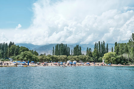 Panoramic view of busy public beach on shores of Issyk-Kul Lake near Cholpon-Ata, Kyrgyzstan. People swimming and relaxing with high Tien Shan mountains and dramatic summer clouds in background.の写真素材