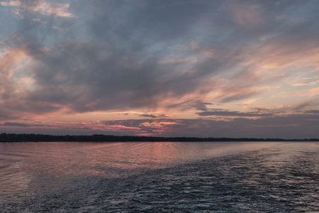 Dramatic sunset over wide Volga River. Sky filled with dark gray and soft pink clouds, their colors reflecting intensely on water s surface, with distant, dark forest silhouette on horizon.の写真素材