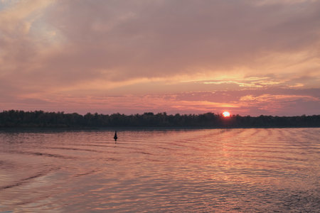 Serene and colorful sunset over Volga River, with sun dipping below dark forest horizon. Sky and water are bathed in soft pink, orange, and red light, creating a peaceful, reflective scene.の写真素材