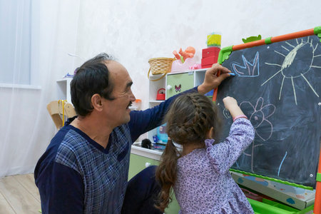 Cheerful grandfather and his young granddaughter are collaborating on drawing on chalkboard in children's room. Girl draws flower while man adds crown. Joyful moment of creativity and learning.の写真素材
