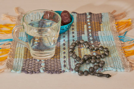 Traditional Iftar components to break fast. Glass of water and dates in turquoise bowl, dark prayer beads or tasbih or misbaha, on brightly colored, handmade woven mat.の写真素材