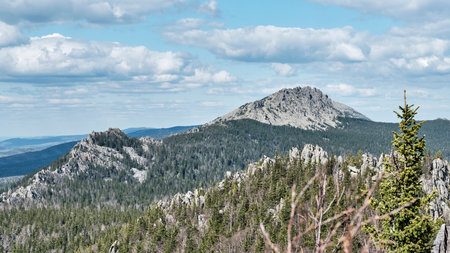 Wide, scenic view across the forested slopes of Taganay National Park, Russia, highlighting the distinct rocky peaks of Mitkinskiye Rocks and distant Otkliknoy Ridge under blue sky with white clouds.の写真素材
