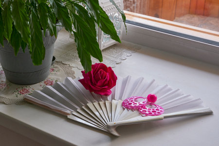 White open folding fan on windowsill with vibrant red rose and large pink statement earrings. Traditional Spanish dance accessories in bright interior setting.の写真素材
