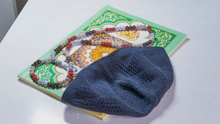 Dark gray knitted kufi hat and prayer beads lying on Arabic language textbook with traditional...